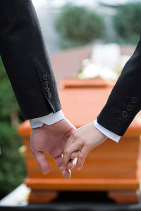 Two people in formal attire holding hands beside an orange casket at a funeral