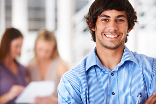 Smiling young man with dark hair wearing a blue striped shirt in a bright office, with two women discussing documents in the background.