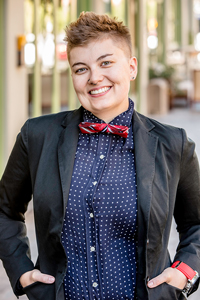 Smiling person with short hair wearing a blazer, polka dot shirt, and red bow tie standing against a blurred urban background.
