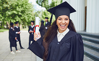 Graduate holding diploma in cap and gown on university campus with other graduates in background