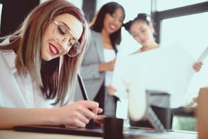 Young female professional using a graphics tablet in an office with two colleagues discussing paperwork in the background.