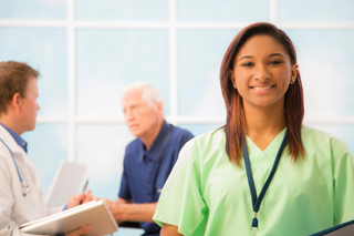 Nurse in green scrubs smiling at the camera with a Doctor and senior patient discussing in the background in a medical office with a bright window