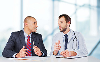 Two professional men in discussion, one wearing business attire and the other in a medical doctor's uniform with a stethoscope, sitting at a table in a bright office setting.