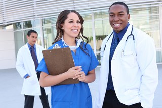 Two medical professionals, a male and a female, smiling and standing outside a hospital, with a male colleague in the background.