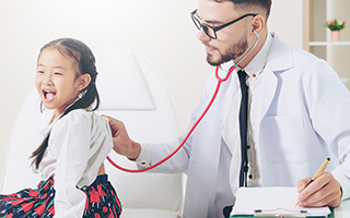 Pediatrician using stethoscope on a young smiling girl in a medical office while taking notes