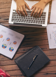 Person typing on laptop with business charts and notebook on wooden desk
