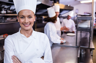Chef in white uniform standing in a commercial kitchen with colleagues working in the background.