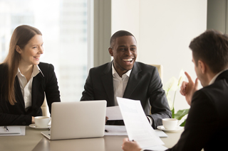 Three business professionals smiling and discussing with documents and a laptop on a desk in a bright office setting.