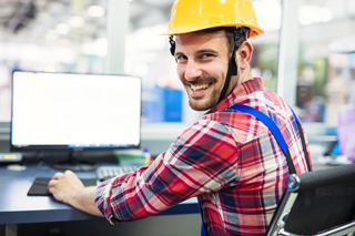 Construction worker in a yellow hard hat sitting at a desk with a computer in an industrial setting