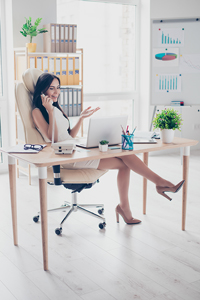 Professional woman sitting at a desk in an office, talking on the phone and smiling, with a laptop and various office supplies around her.