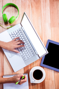 A laptop on a table with green headphones, a coffee, and a notepad around it