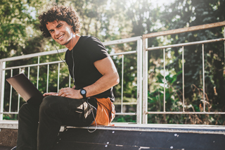 Young man with curly hair sitting outdoors with a laptop on his lap, wearing a black T-shirt and headphones, in a sunny park setting.