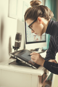 Young woman using a digital tablet with a stylus in a creative home office environment, focusing intently while drawing near a microscope and framed artworks.