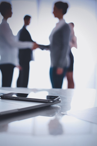 Business professionals shaking hands in background with focus on smartphone and pen on table in foreground in office setting