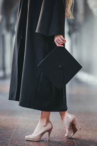 Woman holding graduation cap while wearing black gown and beige high heels