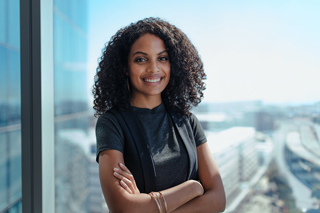 Confident young woman with curly hair smiling in a corporate setting by a glass window with a city view in the background.