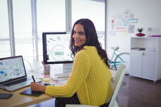 Woman in yellow sweater working creatively with graphic tablet at office desk with dual monitors displaying graphs