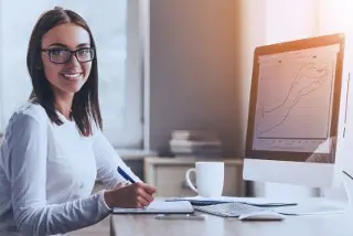 Woman with glasses working at desk with computer and paperwork in office setting