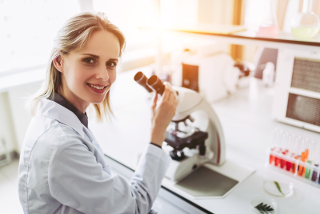 Female scientist using a microscope in a laboratory, smiling at the camera with lab equipment in the background.