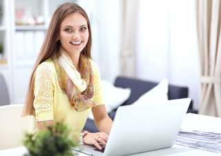 female professional in a yellow sweater and a scarf smiling and working on a laptop