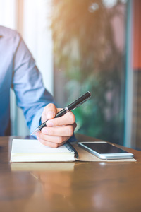 Businessman sitting at a desk writing in a notebook with a pen, smartphone beside him, in an office with natural lighting