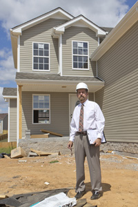 Man in construction hard hat and business attire standing in front of a new two-story house on a construction site.