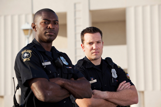 Two police officers standing confidently with arms crossed, wearing uniforms and badges, outside a building.