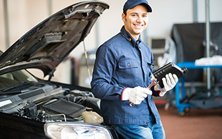 Mechanic in blue uniform holding a car part while standing in front of an open car hood in a workshop.