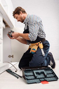 Handyman repairing kitchen sink with tools, wearing plaid shirt and denim jeans with tool belt.