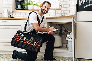 Plumber with tool bag kneeling while fixing sink in a modern kitchen.