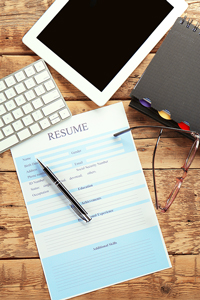 Resume on clipboard with pen, glasses, and electronics on wood desk