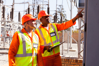 Two electrical engineers in high visibility vests and hard hats inspecting equipment at a power substation, one pointing and explaining, the other holding a clipboard and listening attentively.
