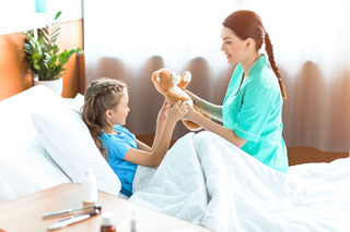 Nurse in teal scrubs giving teddy bear to young girl patient in hospital bed, both smiling in a sunny hospital room with medicines on bedside table.