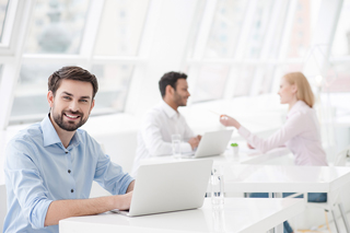 Man with beard working on laptop in bright office space with colleagues discussing in background