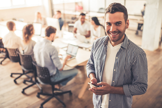 Man with smartphone in an office environment with colleagues working in the background.