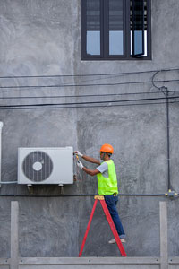 Construction worker in orange helmet using tools to install or repair an outdoor air conditioning unit on a gray building wall, standing on a red ladder.
