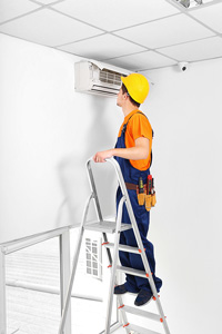 HVAC technician in orange and blue uniform inspecting air conditioner unit on a ladder in an office environment.