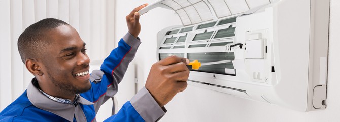 Smiling HVAC technician in a blue and gray uniform working on a wall-mounted air conditioner with a screwdriver