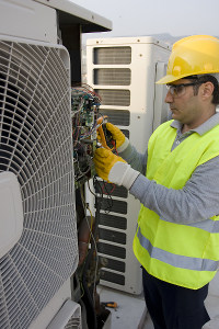 HVAC technician in hard hat and reflective vest using a multimeter to service a large air conditioning unit outdoors