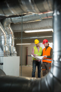 Two HVAC workers with hard hats and high visibility vests discussing over a clipboard in a building under construction, surrounded by exposed ventilation ducts.