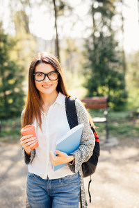 Young woman with glasses holding a coffee cup and folders, wearing a backpack in a park setting.