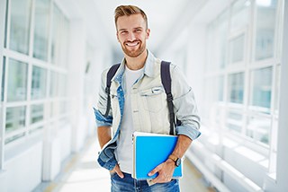 Smiling young male student with beard carrying backpack and blue folder in a bright modern hallway