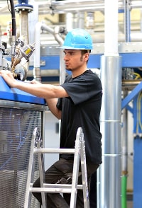 Industrial worker adjusting machinery in a manufacturing plant wearing blue helmet and black t-shirt