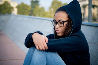 Young woman with glasses and a hooded jacket sitting on a concrete ledge looking thoughtful.