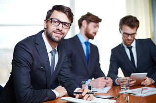 Three professional men in suits at a business meeting, one smiling at the camera while the other two are focused on documents and a laptop.