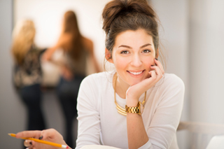 Young woman with a pencil sitting at a table, talking on her mobile phone, with two blurred figures in the background.
