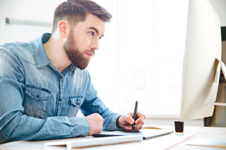 Young man with beard in denim shirt using computer and taking notes in office setting