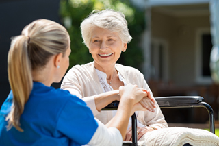 Elderly woman in wheelchair smiling at young nurse outdoors, with hands clasped in a caring gesture.