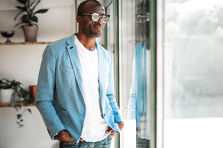 Stylish young man in blue blazer and white t-shirt wearing glasses, standing indoors with a slight smile, looking away from the camera.