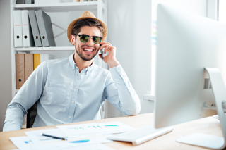 Man in sunglasses and fedora sitting at office desk, working on computer with papers and charts around.